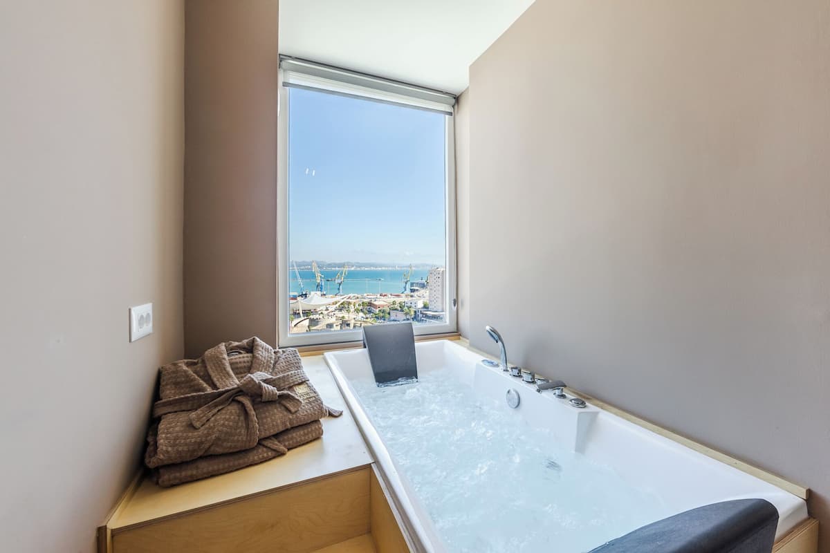 Une salle de bain moderne avec une grande baignoire, une vue sur la mer et une pile d’serviettes sur une étagère en bois.