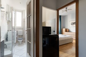 Un bagno moderno con WC, doccia e lavandino. Si vede un letto con materasso bianco e un piccolo comodino. C'è anche un forno a microonde nella stanza.