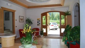 A spacious living area with a wooden ceiling, a dining table, and a red chair. There is a large window with a view of a car and greenery outside. - Hotel Garnì Arnica (Molveno)
