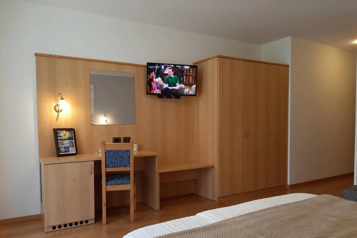 Een slaapkamer met een bed, een houten garderobe, bureau, stoel, televisie en een lijst met een afbeelding aan de muur.