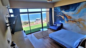 A bedroom with a bed, a chair, a small table, and a large window with a view of the ocean. - WAVES ocean front King Shaka Airport (Desainager)