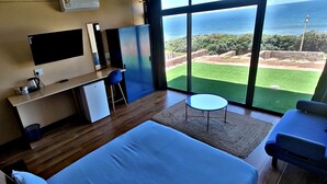 A bedroom with a large bed, a desk with a computer, a chair, and a small table. There is a large window with a view of the ocean and a garden. - WAVES ocean front King Shaka Airport (Desainager)