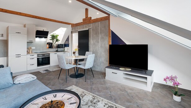 Une cuisine moderne avec des meubles blancs, un coin repas doté d’une table ronde et de chaises, un téléviseur à écran plat posé sur un meuble, et un escalier avec une rampe en verre.