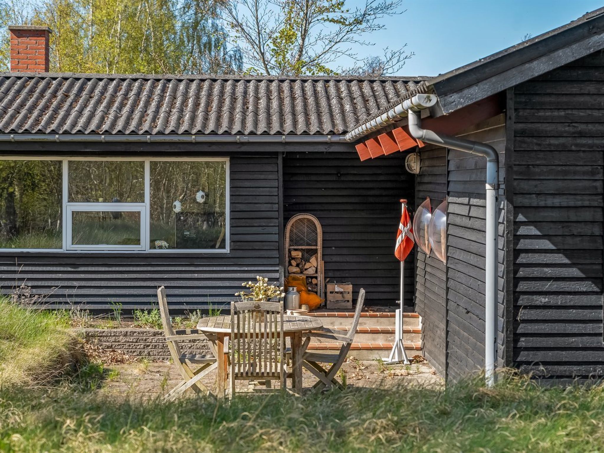 A wooden cabin with a tiled roof, a window, a door, and a small porch with a table and chairs.