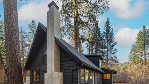 Una cabaña con una chimenea destacada, rodeada de árboles altos y un cielo despejado.