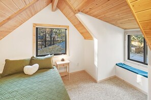 Une chambre avec un plafond en pente, un lit avec une literie verte, une table de chevet, une fenêtre donnant sur des arbres et un banc de fenêtre.