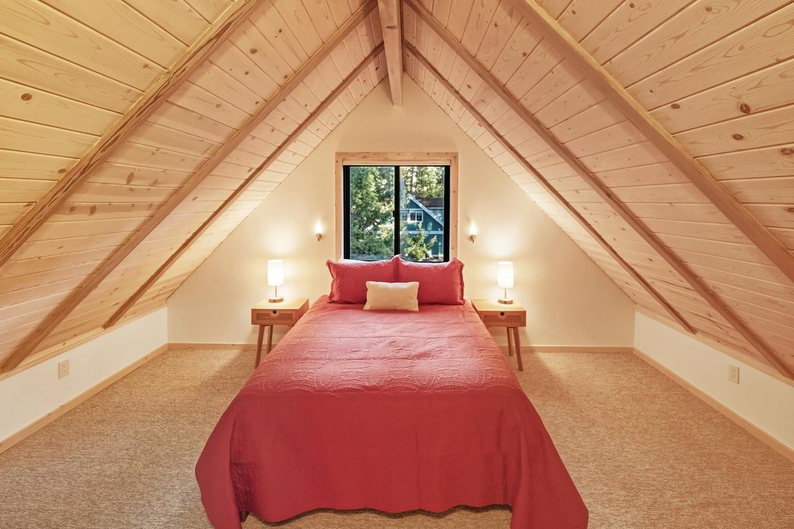 A bedroom with a sloped ceiling, a large window, a bed with red bedding, and bedside lamps.