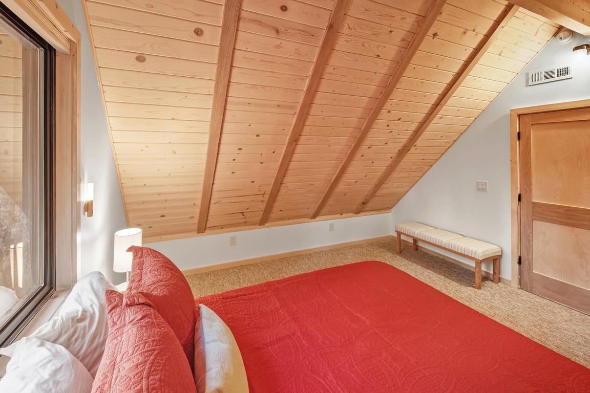 Ein Schlafzimmer mit schrägem Dach, einem Fenster, einem Bett mit roter Bettwäsche, einer Bank und einem Holzschrank.
