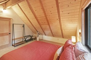 Un dormitorio con techo inclinado, una cama con ropa de cama roja, una ventana con vistas y una puerta de madera.