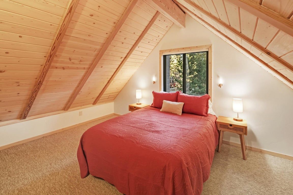A bedroom with a sloped ceiling, a bed with red bedding, two bedside lamps, and a window with a view of trees.