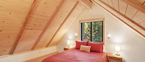 Een slaapkamer met een schuin dak, een bed met rode beddengoed, twee bedlampen en een raam met uitzicht op bomen.