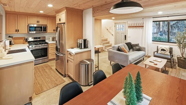 Una cocina moderna con electrodomésticos de acero inoxidable, una zona de comedor con mesa de madera y un espacio de sala de estar acogedor con sofá y una escalera que sube a otro nivel.