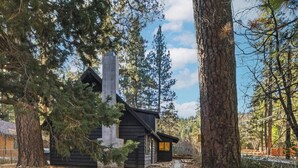 Una cabaña rústica con chimenea de piedra, rodeada de árboles altos y un camino de piedra que lleva a la entrada.