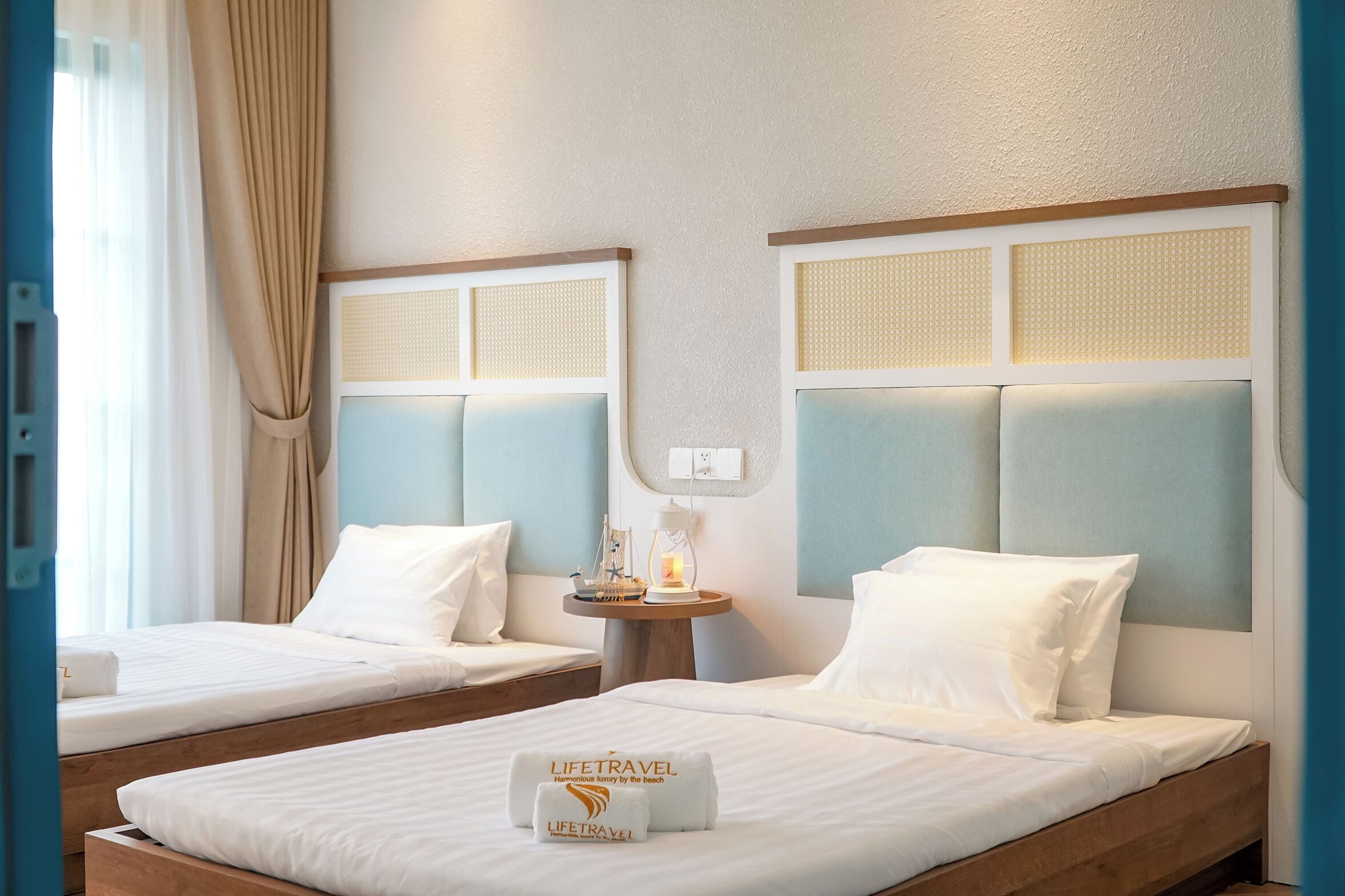 A hotel room with two beds, each with a headboard, a small table with a lamp, and a towel with the hotel name.