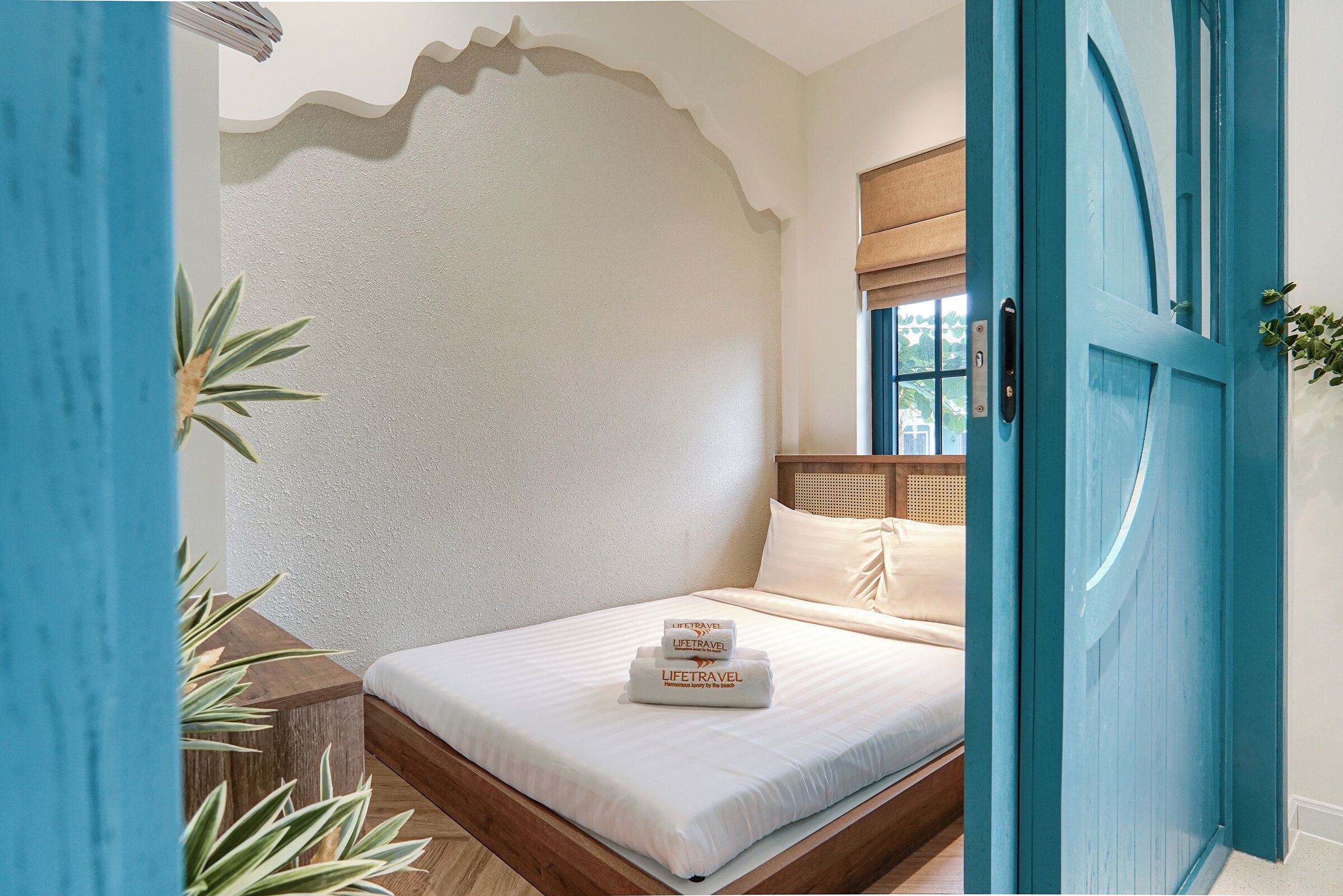 Una cama bien hecha con edredón blanco y dos toallas apiladas encima, cabecera de madera, ventana con persiana de madera y una puerta azul.