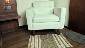 Ein weißer Sessel steht in einer Ecke des Raumes mit Holzboden, einem kleinen Teppich und einem Holztischchen mit einer Lampe.