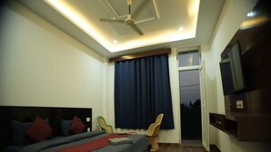 A bedroom with a bed, a ceiling fan, a TV, a window with curtains, and a door leading outside. - The Jungle Hut (Dharamshala)