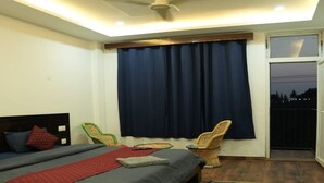 A bedroom with a bed, a ceiling fan, a window with curtains, a chair, and a balcony. - The Jungle Hut (Dharamshala)