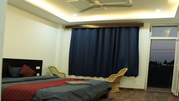 A bedroom with a bed, a chair, a window with curtains, and a ceiling fan. - The Jungle Hut (Dharamshala)