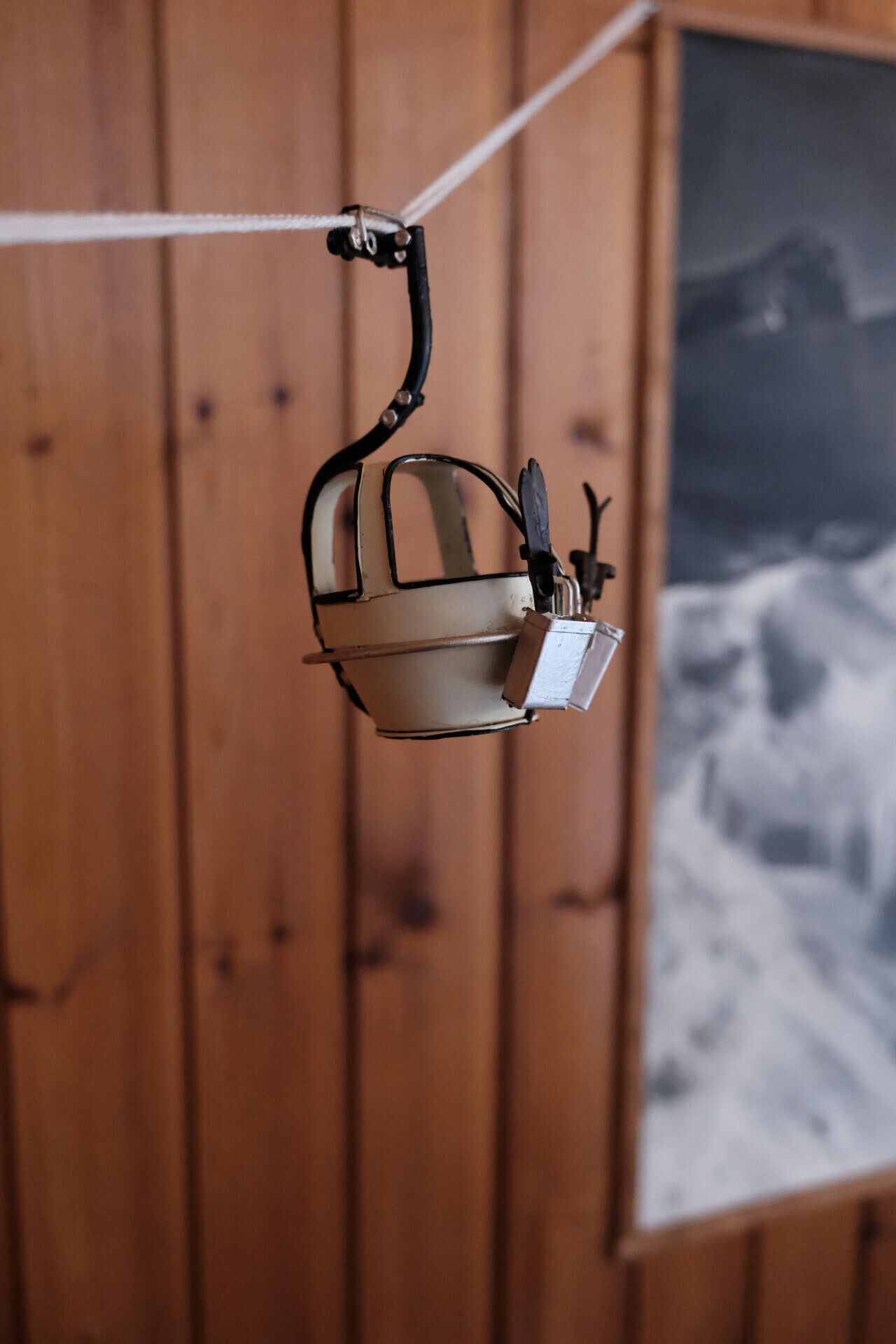 Un objeto parecido a una cesta metálica colgando de un cable, con un fondo de madera y una imagen borrosa de una montaña nevada.