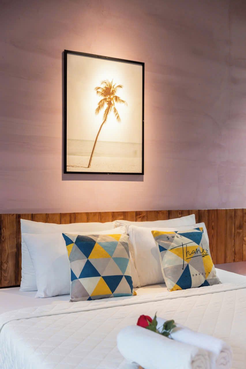 Una cama bien hecha con edredón blanco y almohadas estampadas, un cuadro enmarcado con una palmera en la pared y un cabecero de madera.