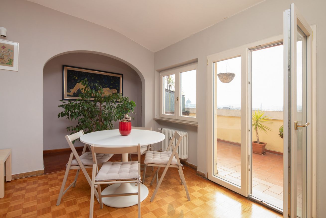 A room with a round table, chairs, a potted plant, a painting, and a view through a glass door.