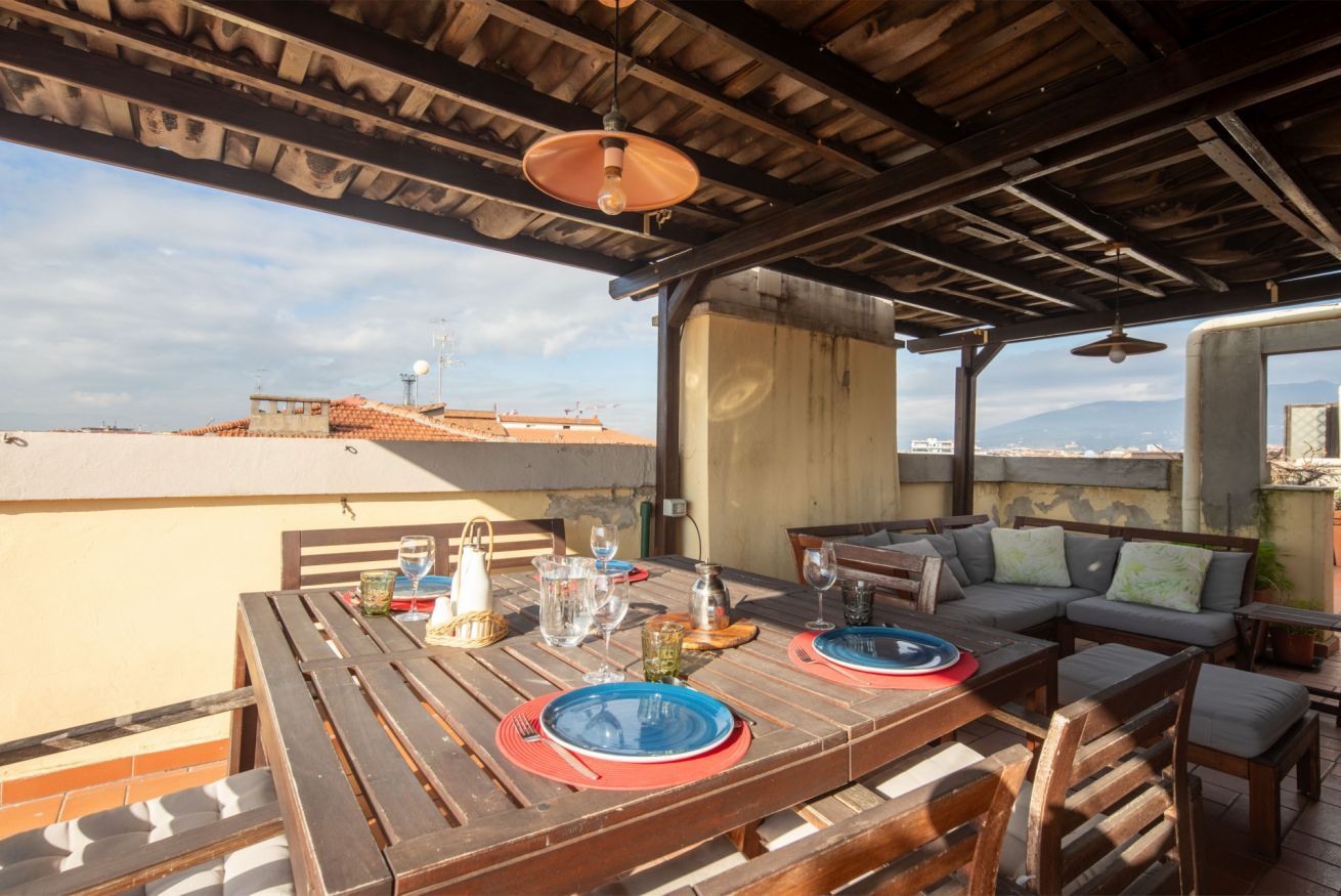 An outdoor patio with a wooden table set for two, a sofa, and a view of the cityscape.