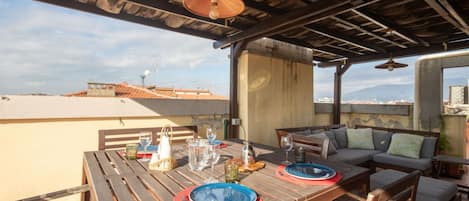 An outdoor patio with a wooden table set for two, a sofa, and a view of the cityscape.