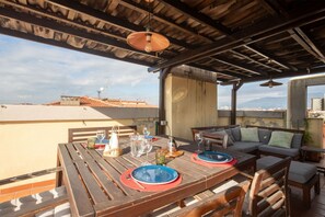 An outdoor patio with a wooden table set for two, a sofa, and a view of the cityscape.