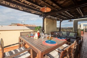 An outdoor patio with a wooden table set for two, a sofa, and a hanging light fixture.