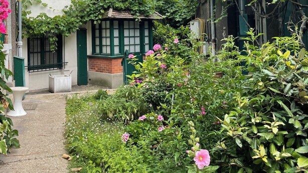 En hvid bygning med grønne skodder, en murstensdel og en have med lyserøde blomster.