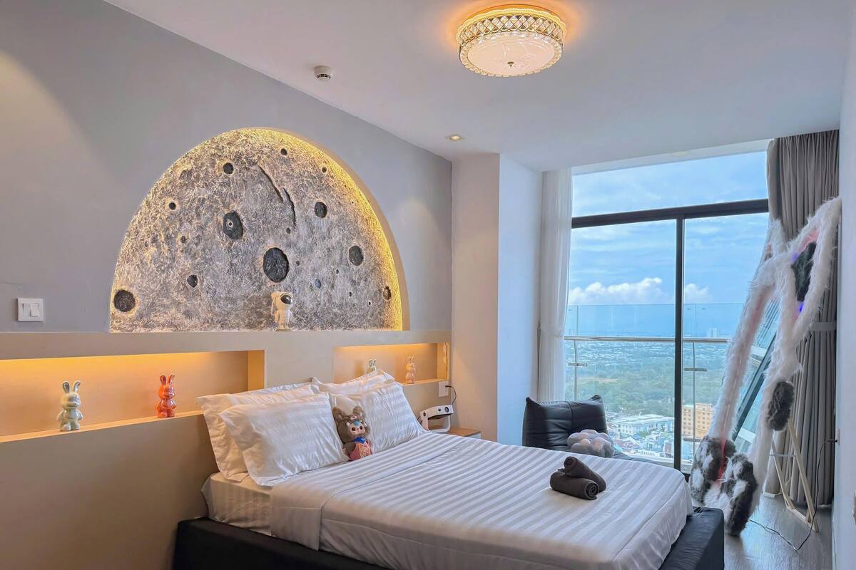 A modern hotel room with a large bed, a wall mural of the moon, a view of the city, and a balcony with a railing.