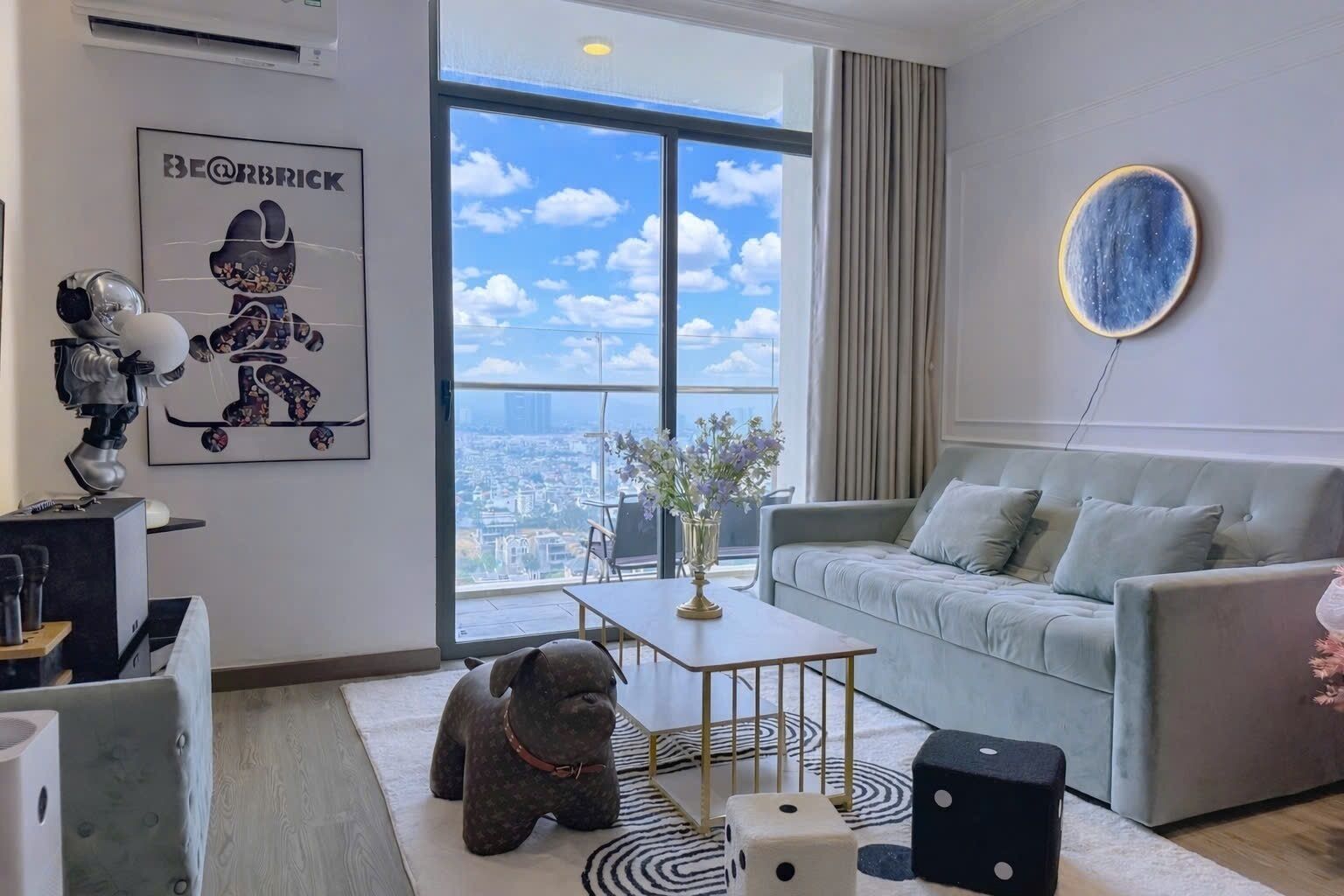 A modern living room with a grey sofa, a coffee table, a dog statue, and a city view through large windows.