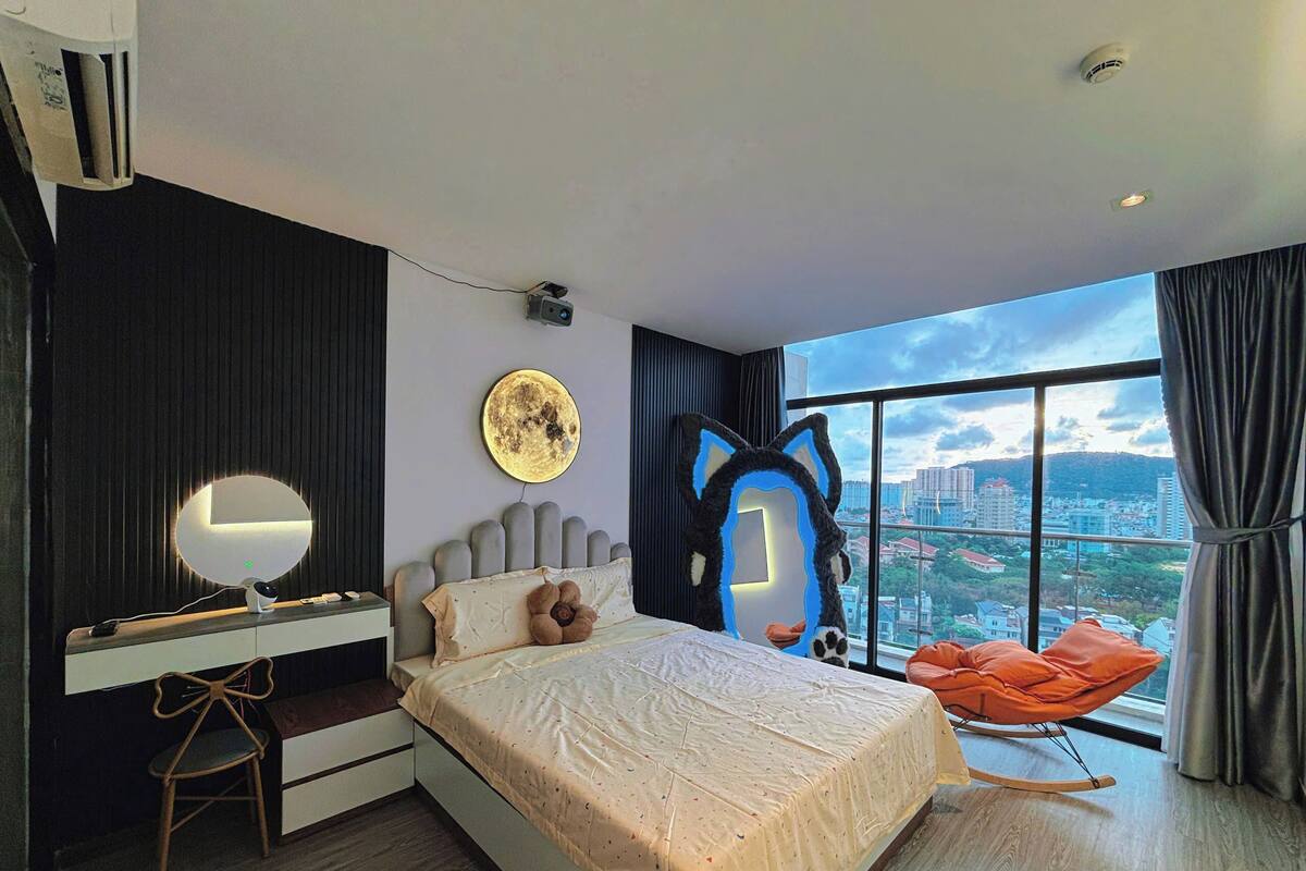 A modern bedroom with a large bed, a desk with a mirror, a chair, and a view of a cityscape through a large window.