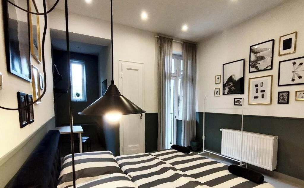 A modern bedroom with a bed, bedside table, and a hanging lamp. There is a window with curtains, a radiator, and framed pictures on the wall.