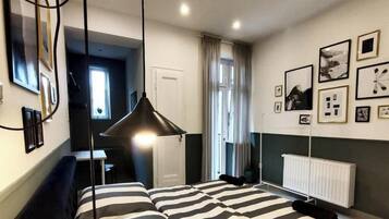 A modern bedroom with a bed, bedside table, and a hanging lamp. There is a window with curtains, a radiator, and framed pictures on the wall.