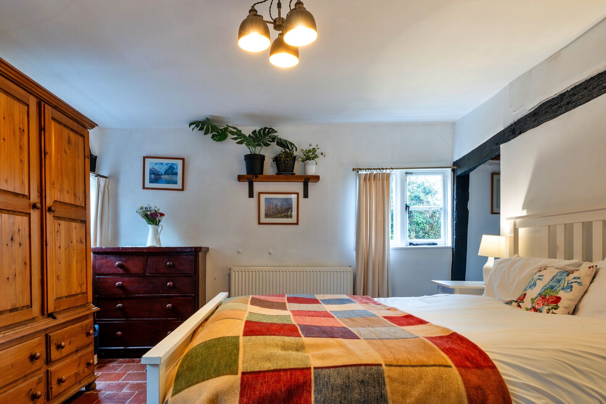 Een slaapkamer met een bed, een houten garderobe, een dressoir, planken met planten, een raam en een plafondlamp.
