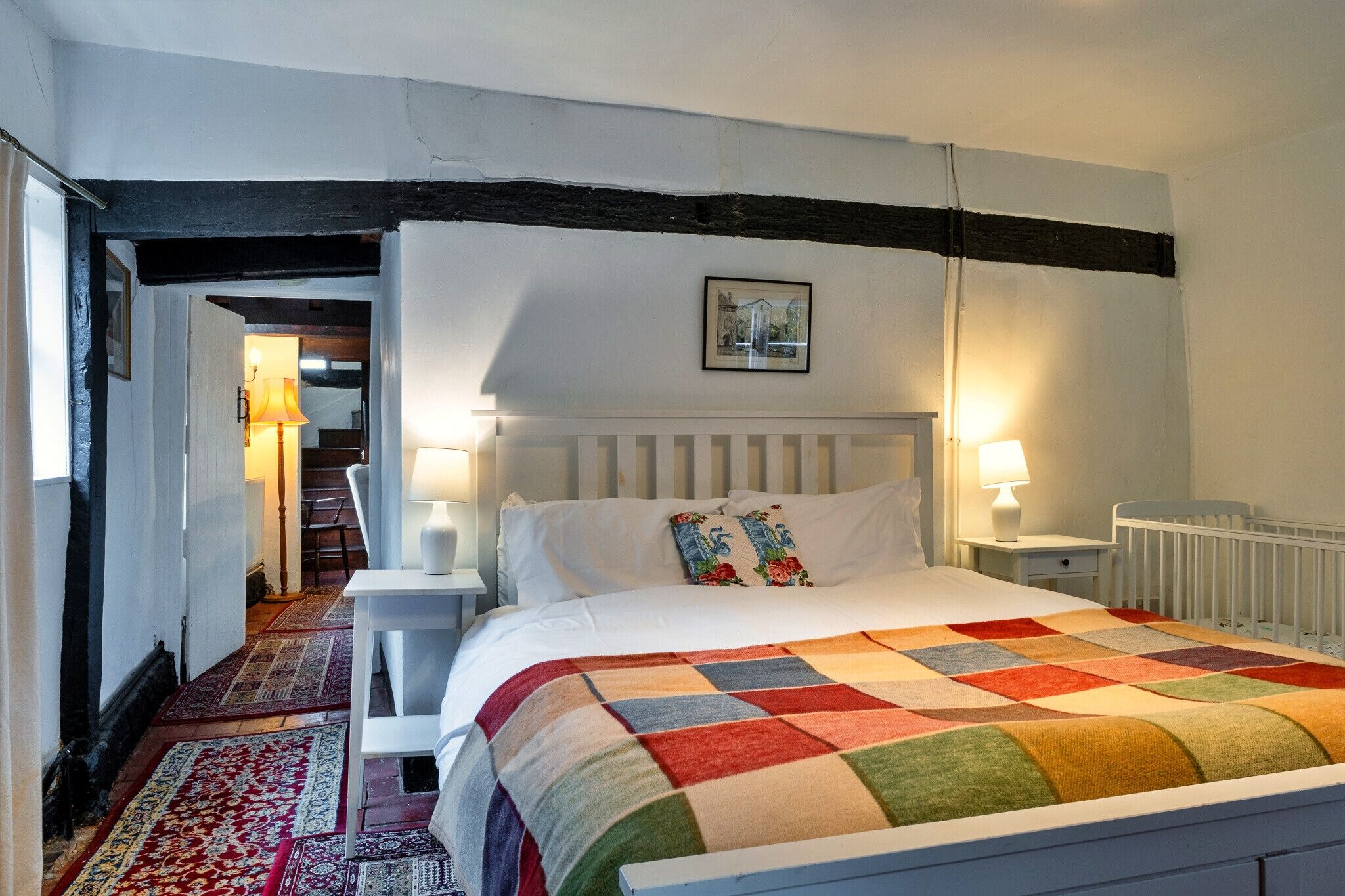 A bedroom with a white bed, colorful quilt, white nightstands, a white chair, a red and white patterned rug, and a black beam on the ceiling.