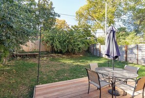En terrasse med et rundt bord og stole, omgivet af en træterrasse og en græsplæne med træer.