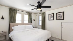 Ein Schlafzimmer mit einem Bett, einem Deckenventilator, einem Nachttisch, einer Lampe und zwei mit Vorhängen versehenen Fenstern.