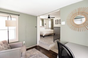 Een slaapkamer met een bed, een bureau, een stoel, een raam met zonwering, een lamp, een plafondventilator en een spiegel aan de muur.
