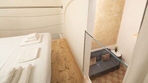 Ein Schlafzimmer mit Bett, Holzboden, einer Glaskabine und einer kleinen Sitzecke mit Sofa und Kissen.