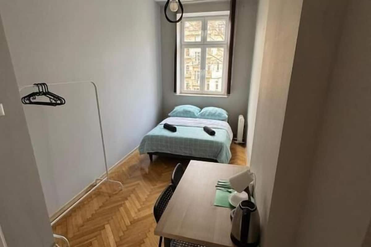 A small, well-lit room with a wooden herringbone floor, a single bed with a blue bedspread, a table with a chair, and a hanging light fixture.