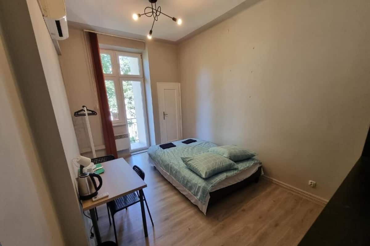 A small, well-lit room with a bed, a table with a kettle, and a chair. There is a door leading to a balcony with a view of trees.
