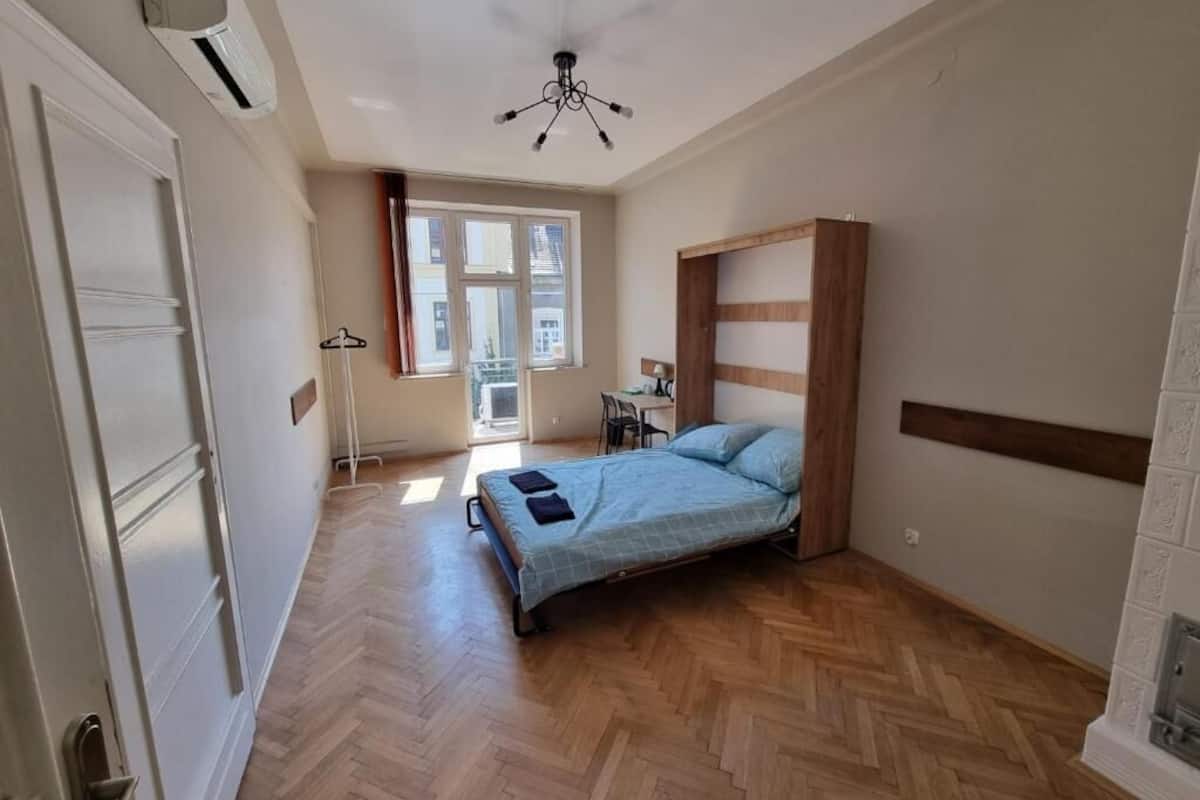 A bedroom with a bed, wooden floor, a ceiling fan, and a window with curtains.