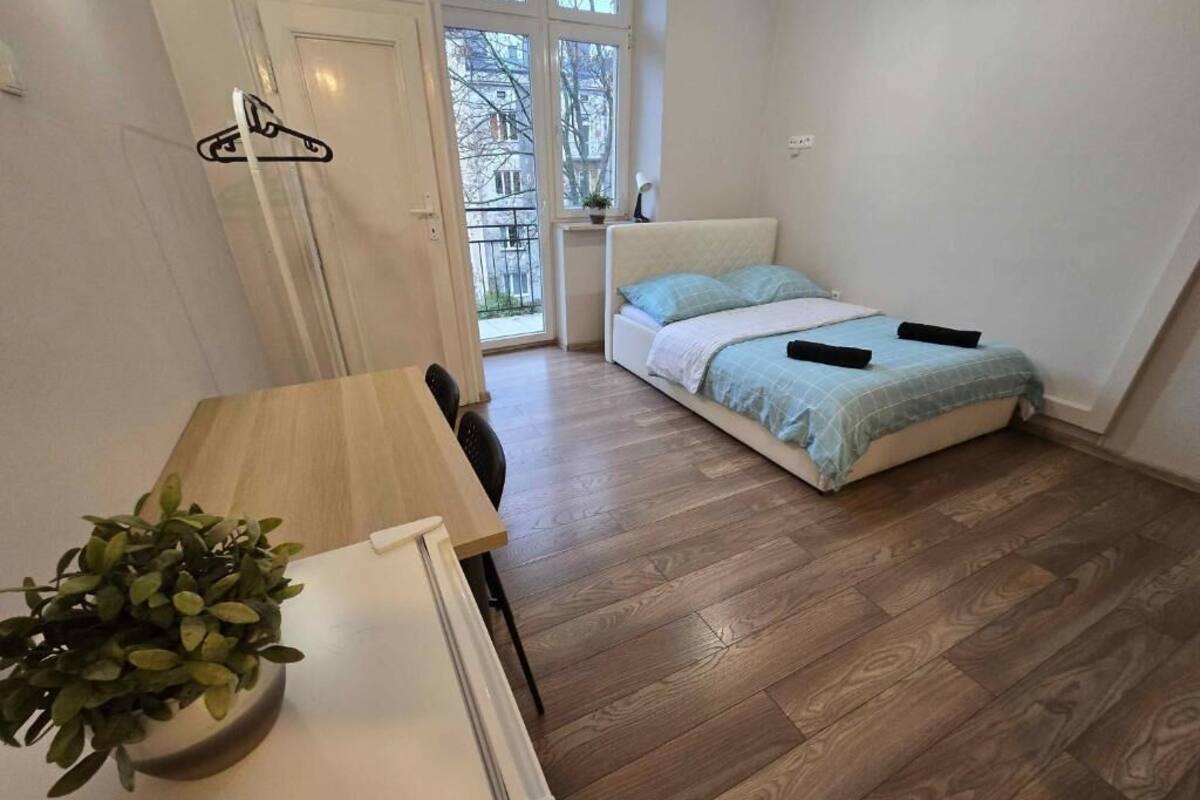 A small, tidy room with a bed, a table with a plant, and a window with a view of buildings.