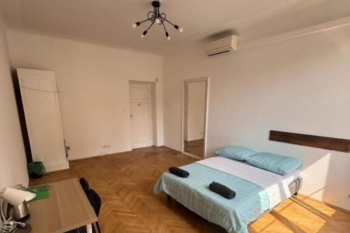 A room with a bed, a desk, and a chair. There is a door and an air conditioning unit on the wall.