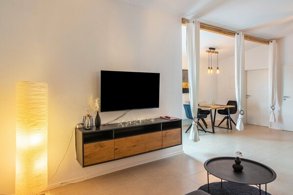A modern living room with a flat-screen TV mounted on a wooden console, a round coffee table, and a dining area with a table and chairs.