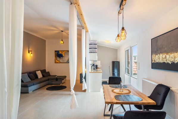 A modern living room with a sofa, a dining table with chairs, a kitchen area, and a large window.