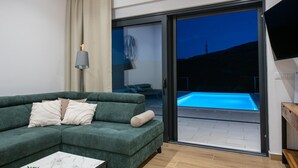 Una sala de estar moderna con un sofá verde, una mesa de centro de mármol, un televisor y un ventanal corredero que da a una zona de piscina exterior.